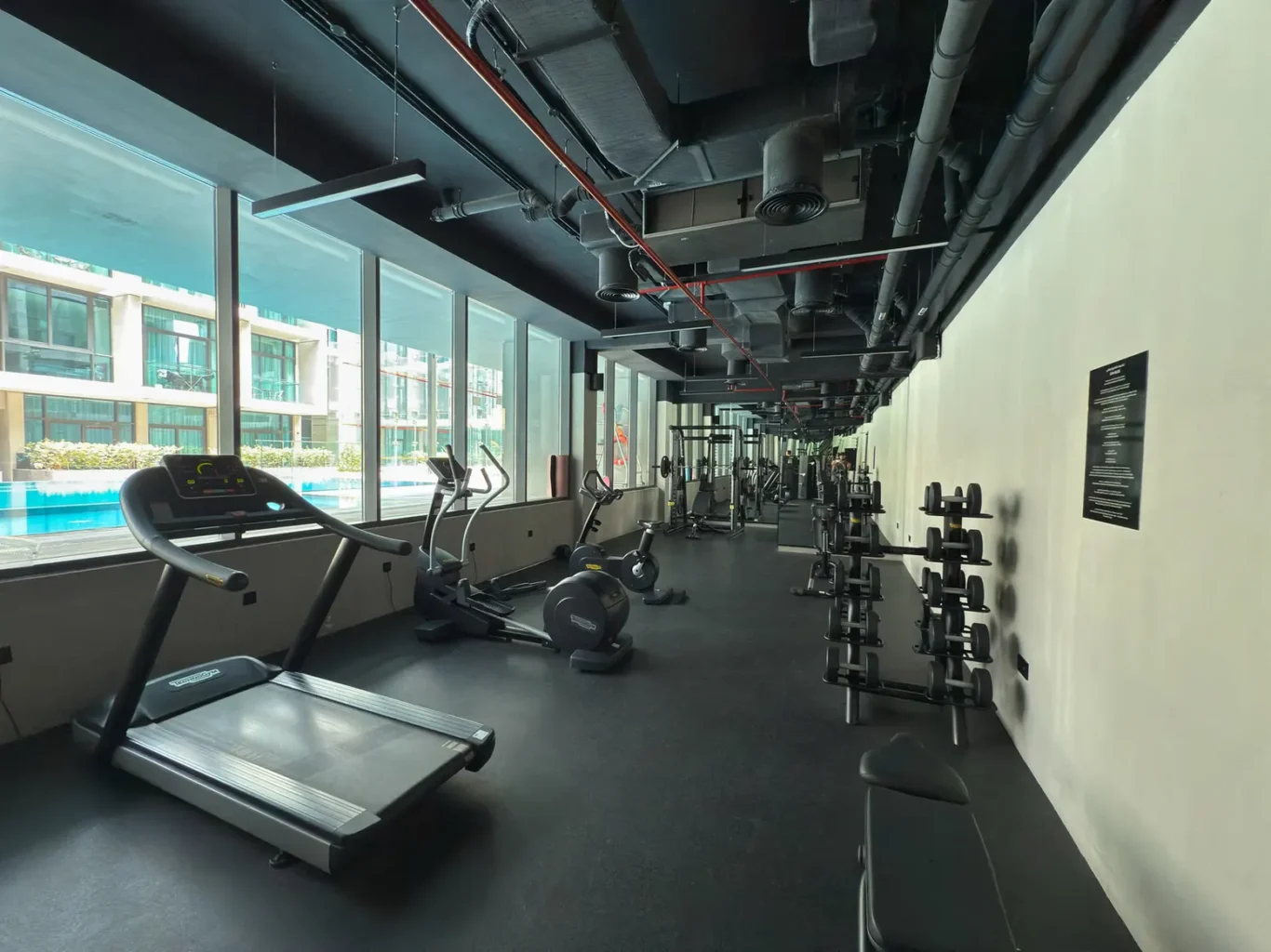 Sunlit gym interior with treadmills and cardio machines along large windows facing an outdoor pool area.
