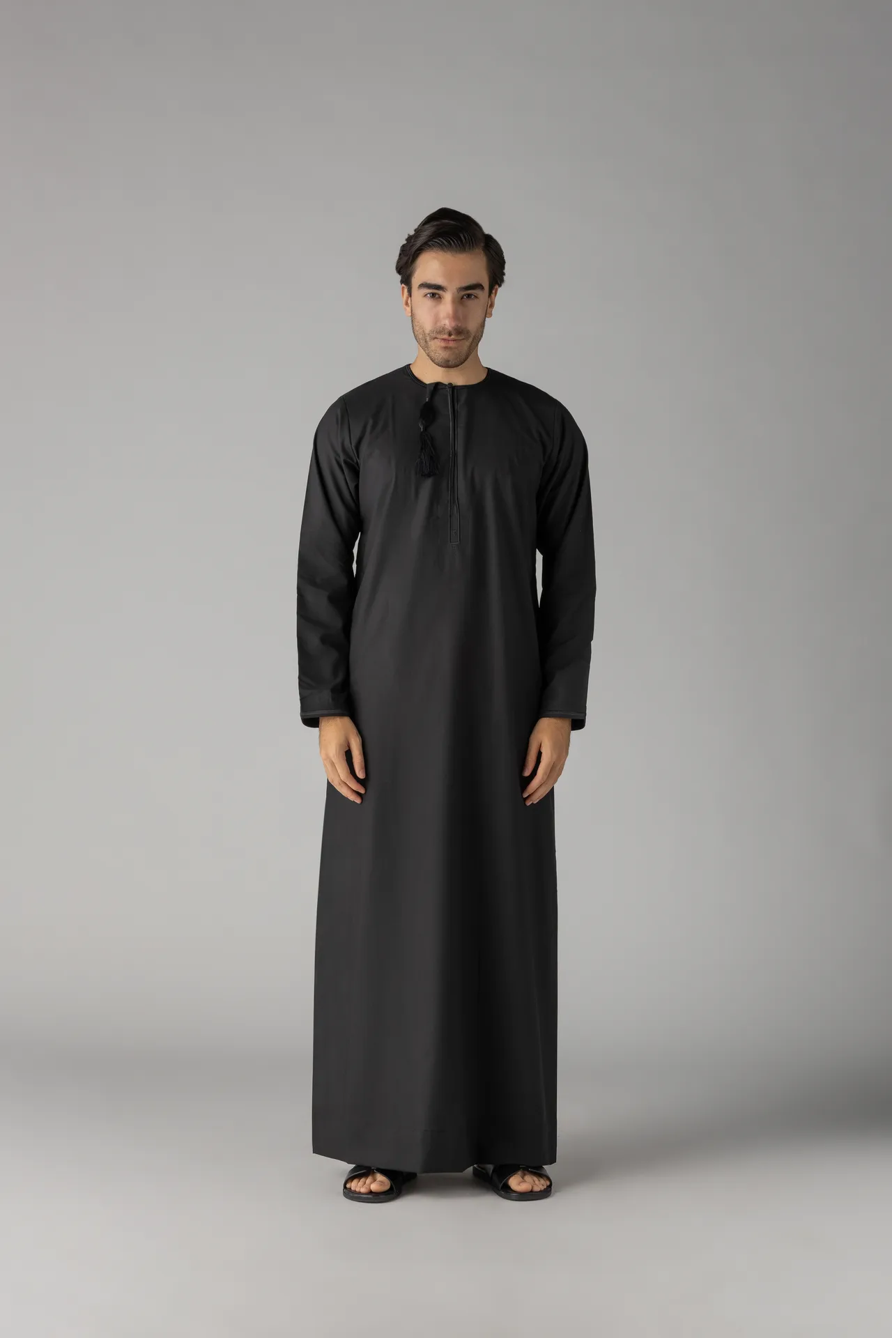 Man wearing a long black thobe, standing against a gray backdrop, hands at sides, sandals visible at feet