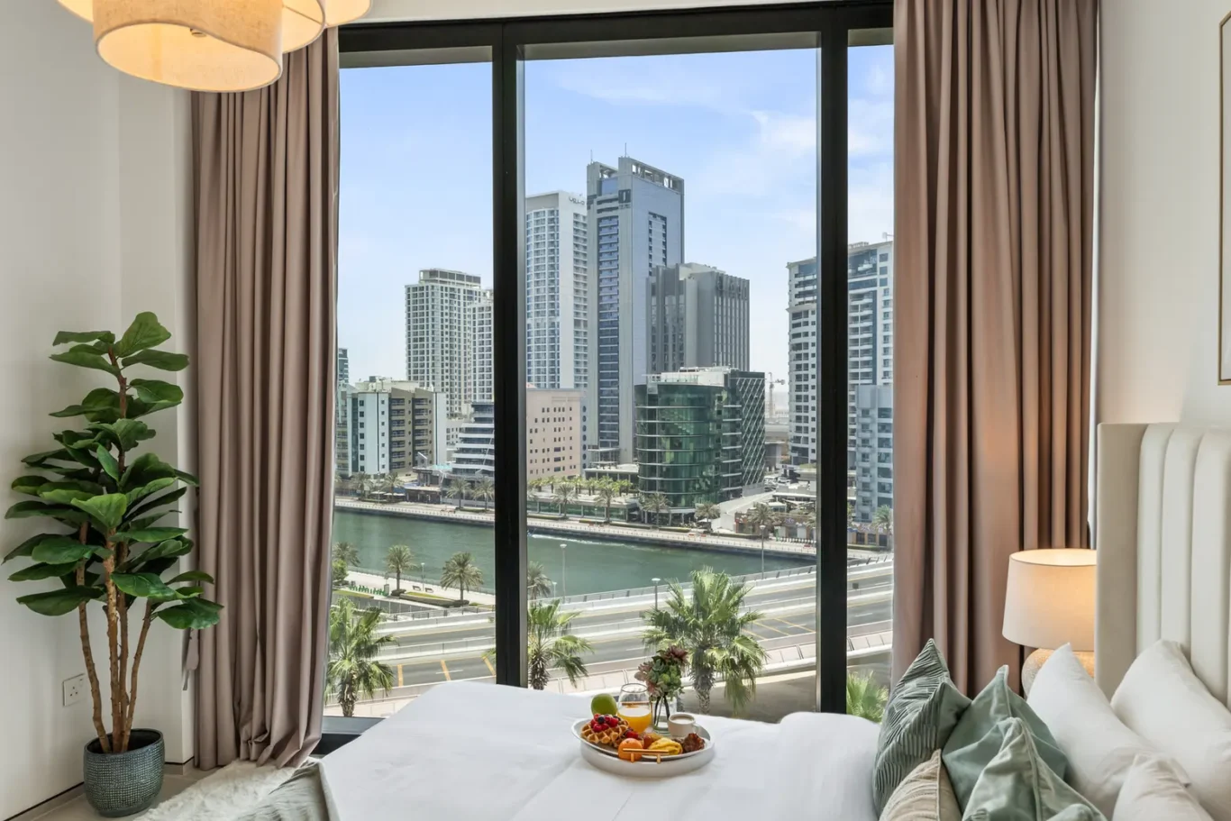 Hotel room with a large window overlooking a city skyline, bed with breakfast plate and greenery nearby.