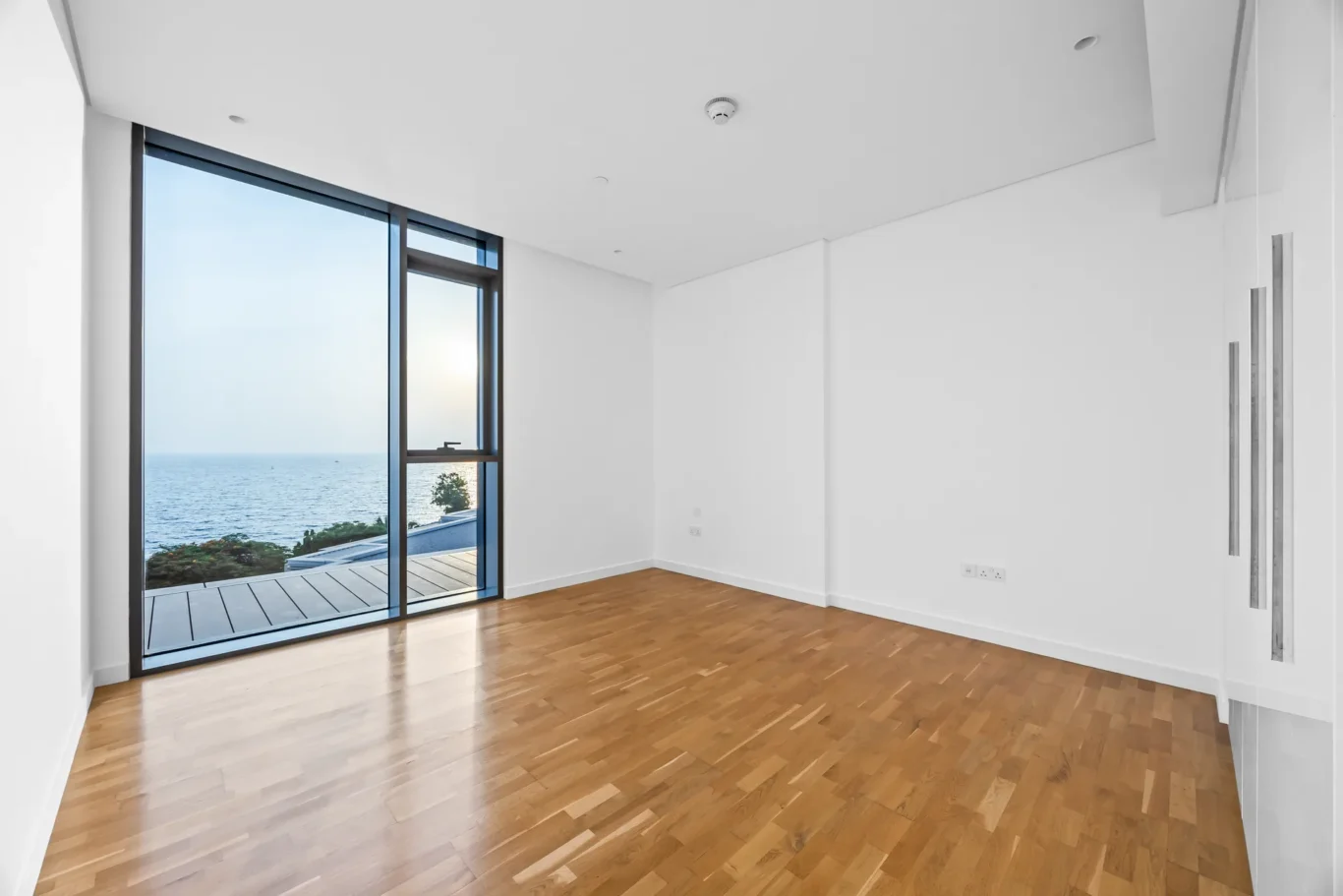 Bright empty room with wood floor and a large sliding glass door opening to a deck overlooking the ocean.
