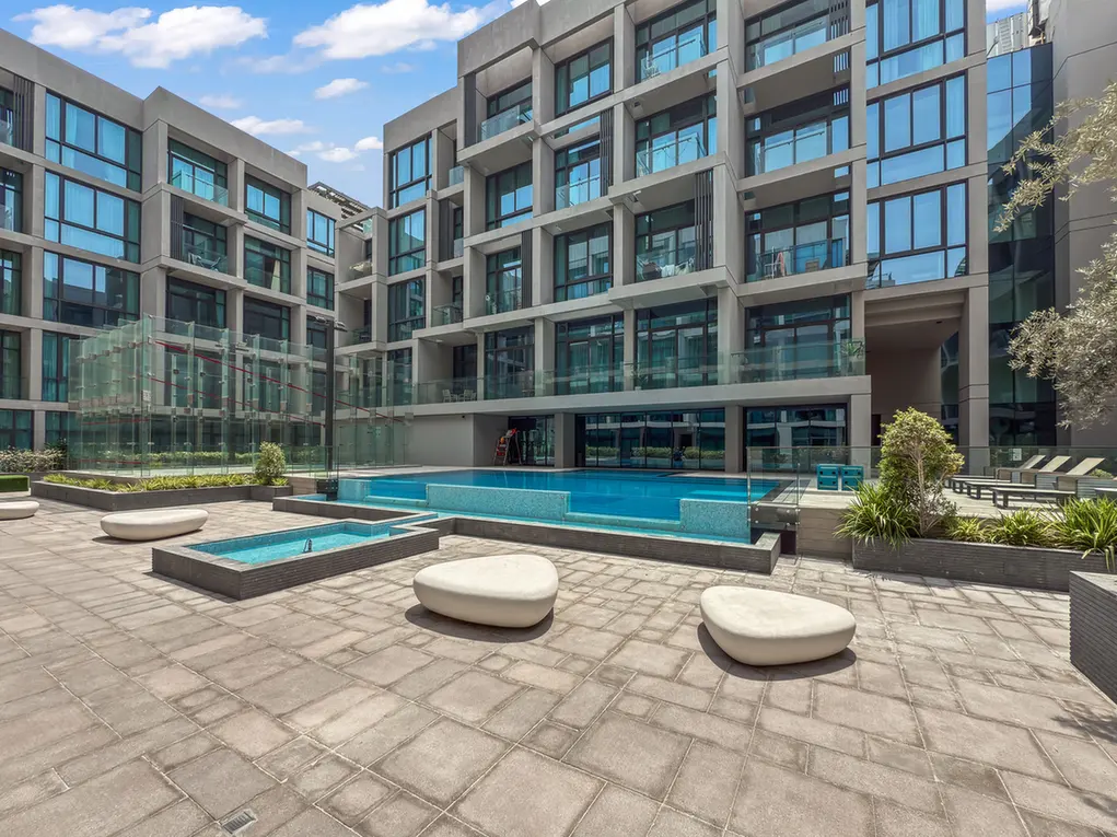 Modern courtyard of a multi-story residential building with glass balconies and a blue-tiled pool area under a sunny sky