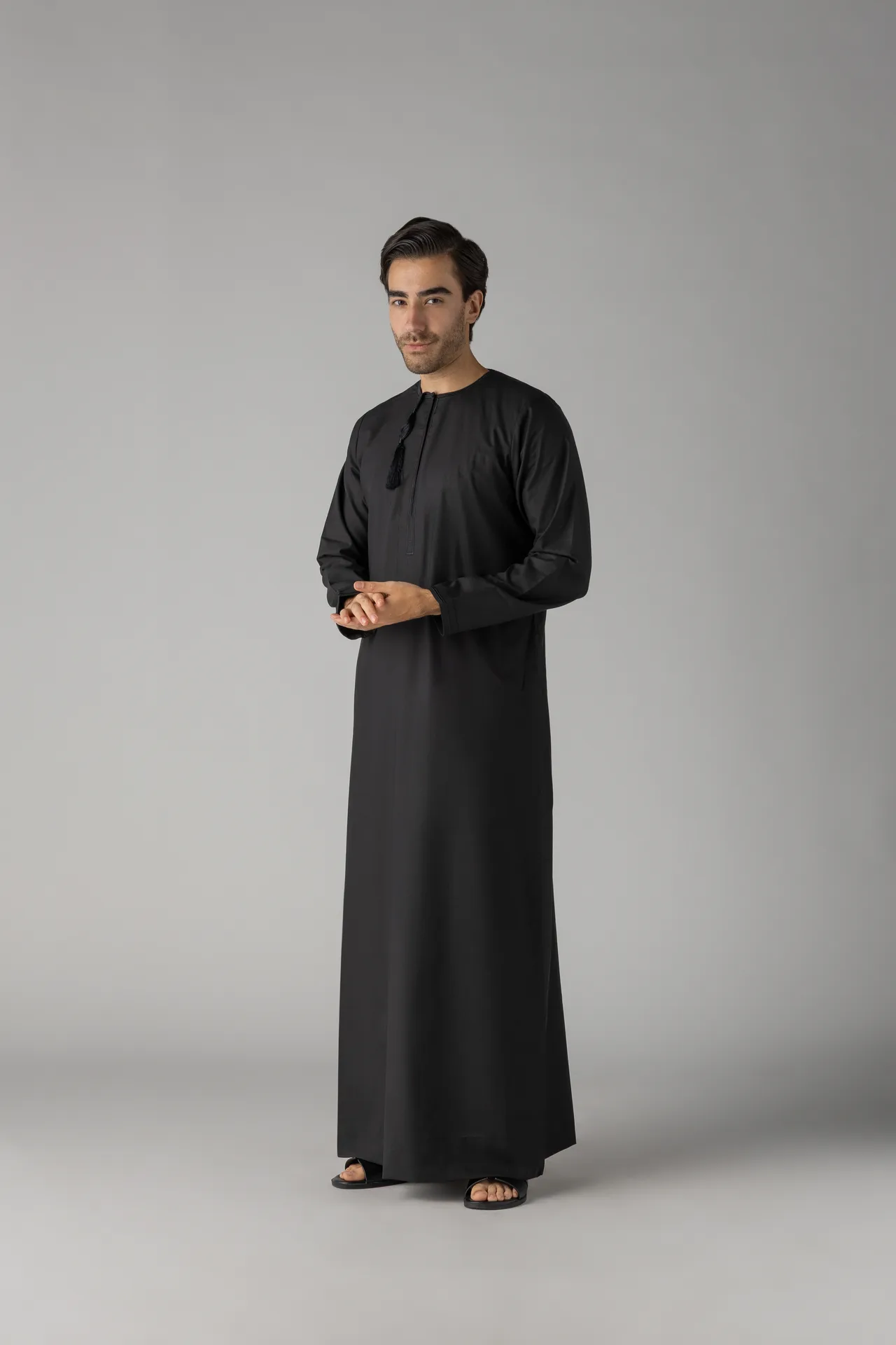 Full-length photo of a man in a black traditional thobe standing against a neutral gray studio backdrop, hands lightly clasped in front, wearing black sandals.