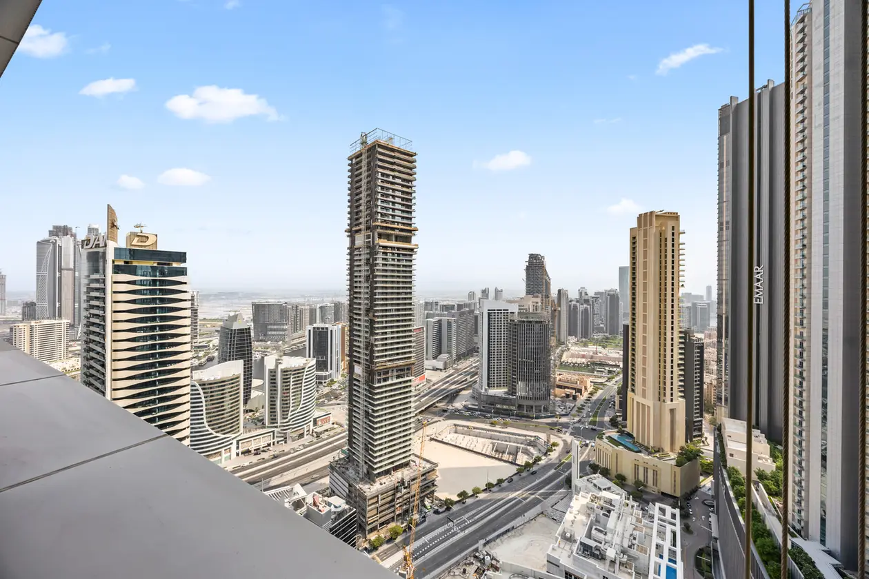 City skyline with numerous skyscrapers, including a tall under-construction tower, beside a multi-lane roadway under a blue sky.