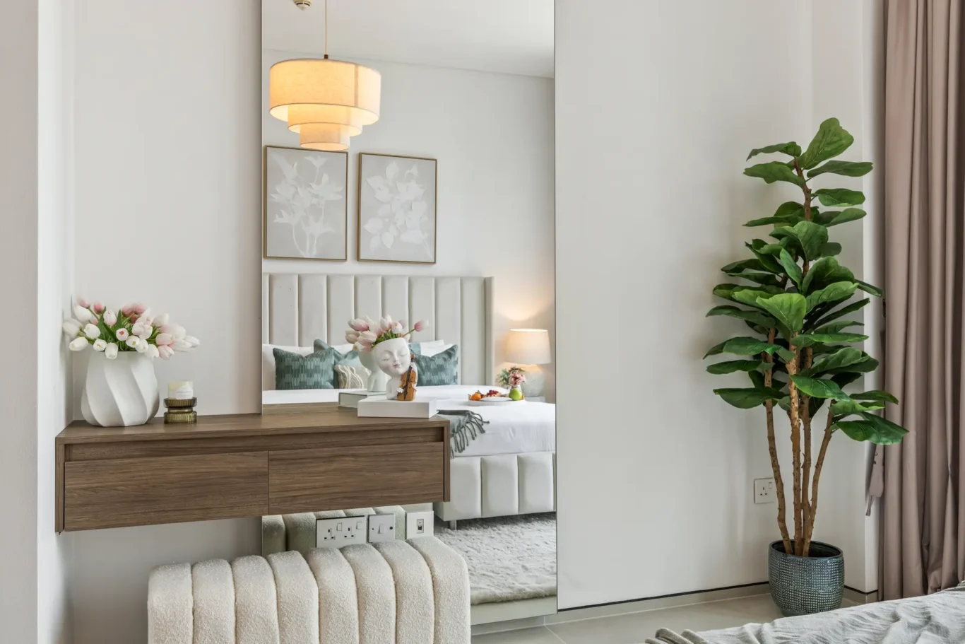 Bedroom interior with a floating wooden dresser, tufted headboard bed, and framed botanical art reflected in a large mirror beside a vase of pink and white tulips.