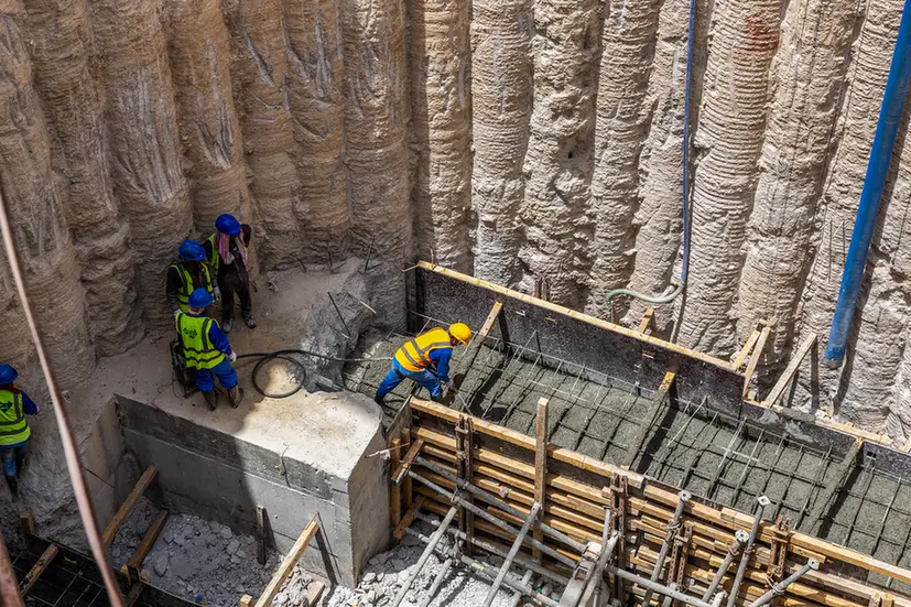 Construction workers in safety vests and helmets pour concrete into wooden formwork at a deep excavation with rocky walls nearby.