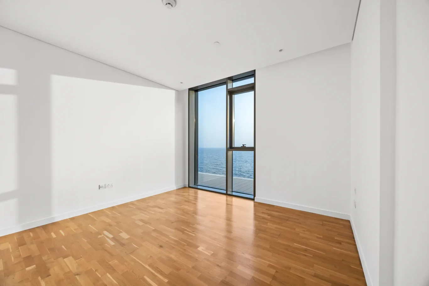 Bright empty room with light wood floor and large glass door overlooking the sea. Close-up shows white walls and an ocean view through the door.
