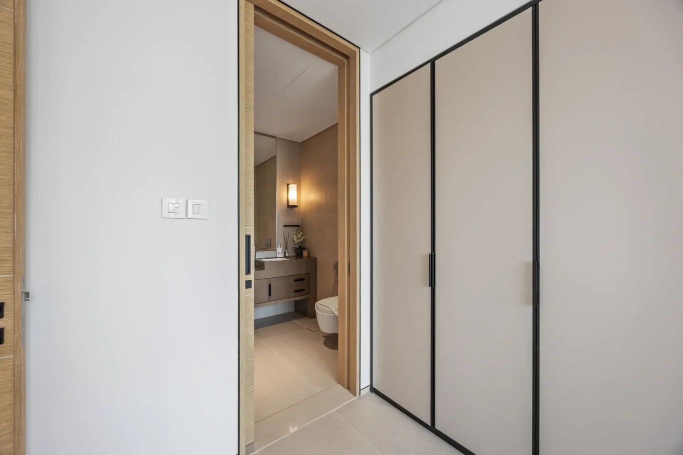 Open doorway into a modern bathroom; beige tiles, wooden vanity, sink, mirror, toilet, and warm wall light.