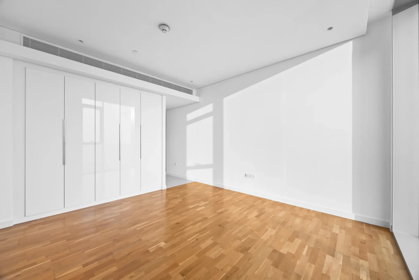 Empty modern room with white built-in wardrobes along the left wall, hardwood floor, and bright natural light from windows.