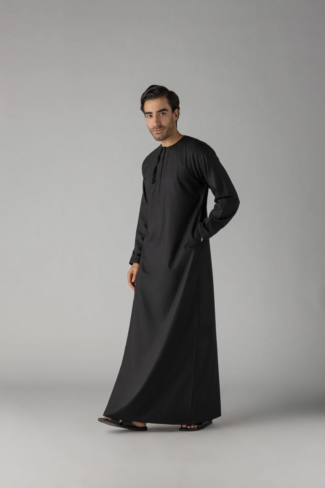 Man wearing a long black traditional robe (abaya) standing with one hand in his pocket against a gray studio backdrop.