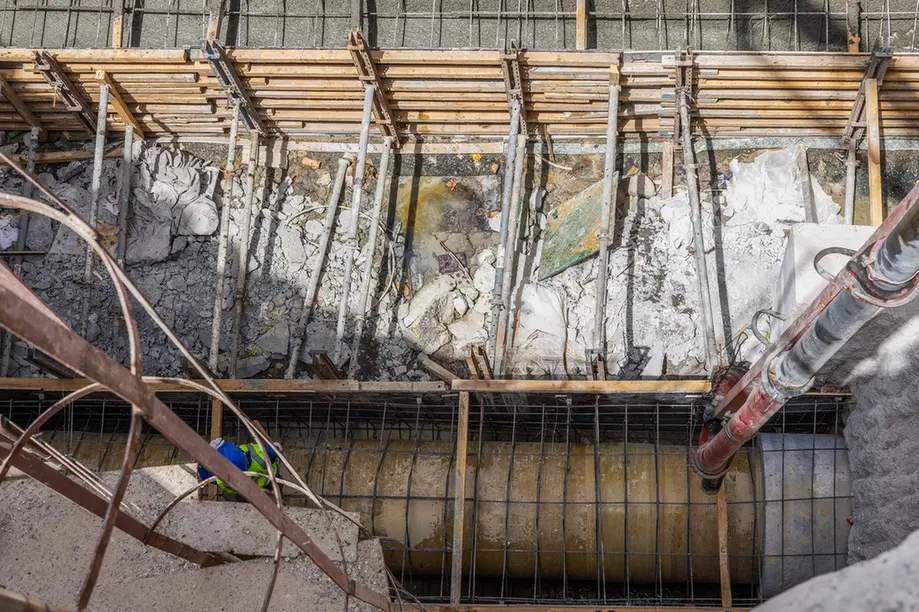 Worker in safety gear stands at the edge of a deep construction trench with exposed rebar and wooden formwork above.