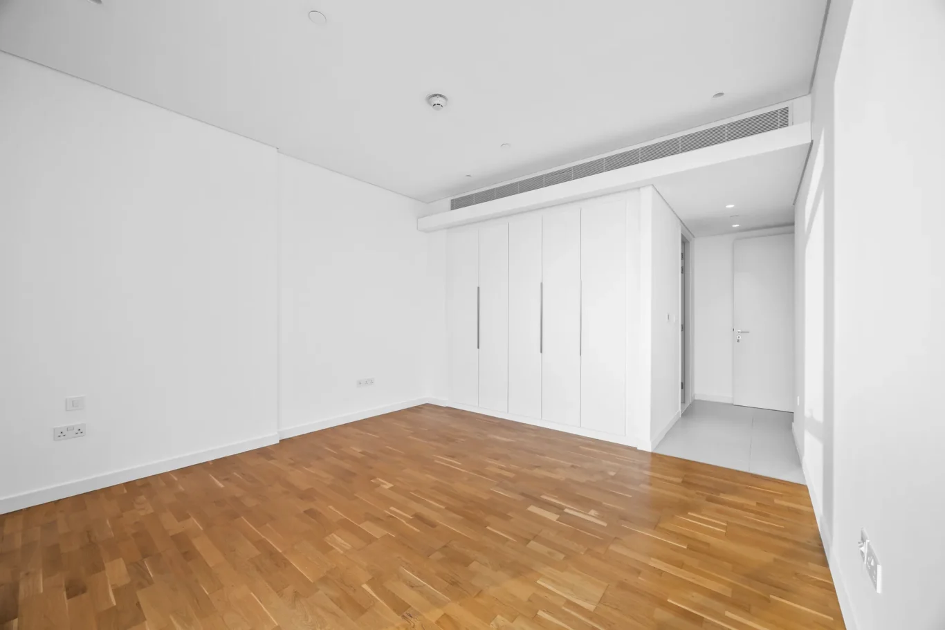 Bright, minimalist room with white walls and a built-in white wardrobe along one wall; hardwood floor and ceiling vents visible.