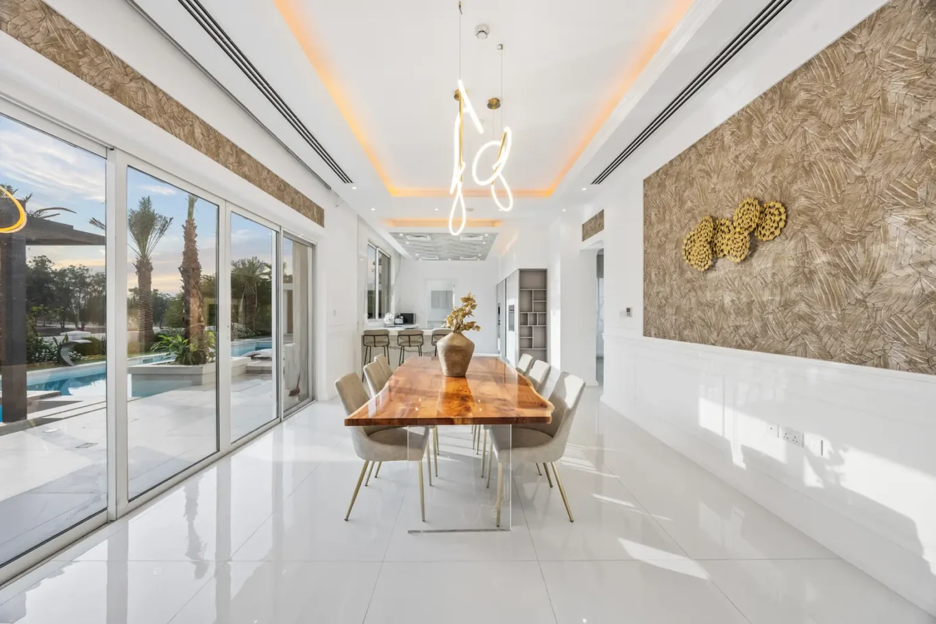 Bright, modern dining room with a long wooden table and beige chairs, overlooking a pool area through large glass sliding doors.