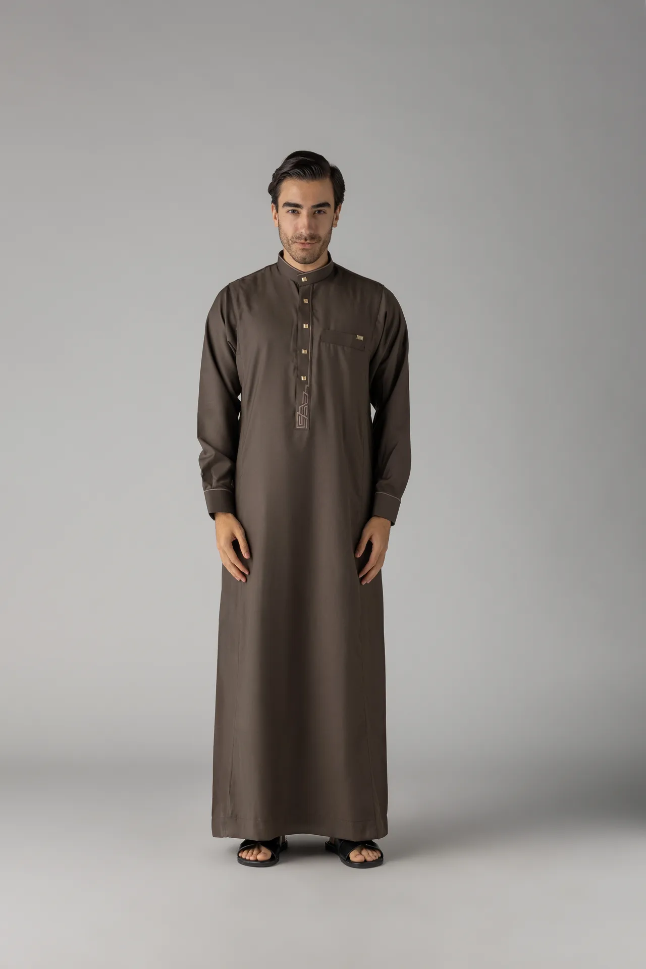 Man wearing a brown traditional thobe with gold buttons, standing against a gray studio backdrop, wearing black sandals.