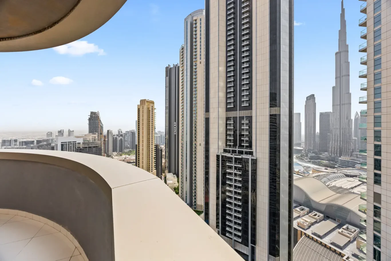 Curved balcony railing in the foreground overlooks a cluster of tall glass skyscrapers under a clear blue sky (city skyline).