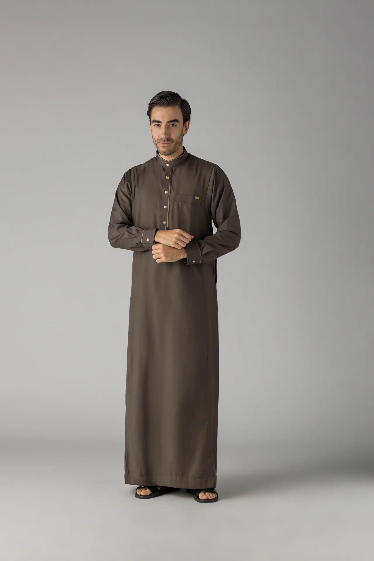 Man wearing a long brown traditional thobe, standing against a neutral gray background with hands folded at the waist and sandals visible.