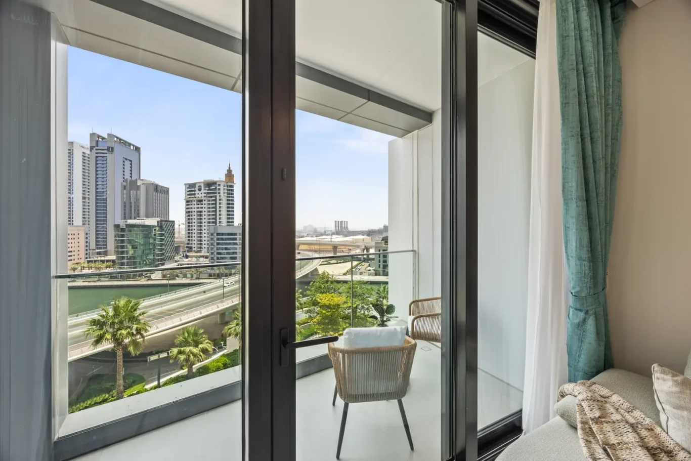 Balcony seating with two wicker chairs overlooking a city skyline and waterway beyond the glass doors, with teal curtains framing the view.