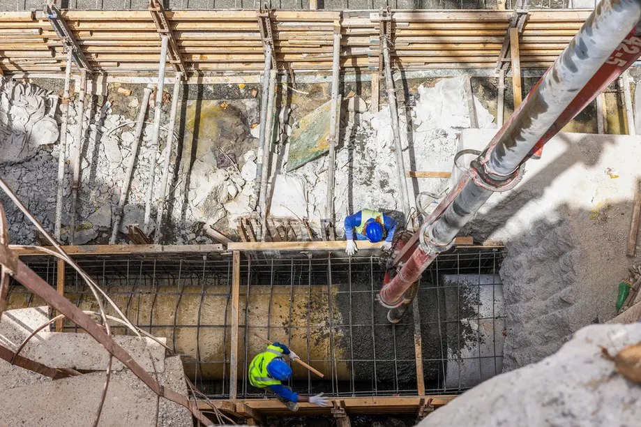 Aerial view of a construction site: two workers in blue helmets and neon vests on a wooden scaffold above a deep rectangular trench with steel reinforcement bars.
