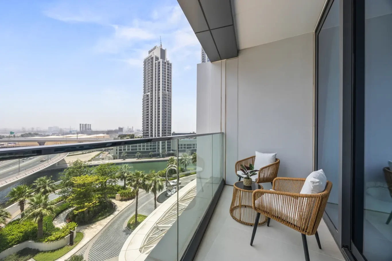 Balcony seating with two rattan chairs and a small table overlooking a cityscape with palm trees and a road below.