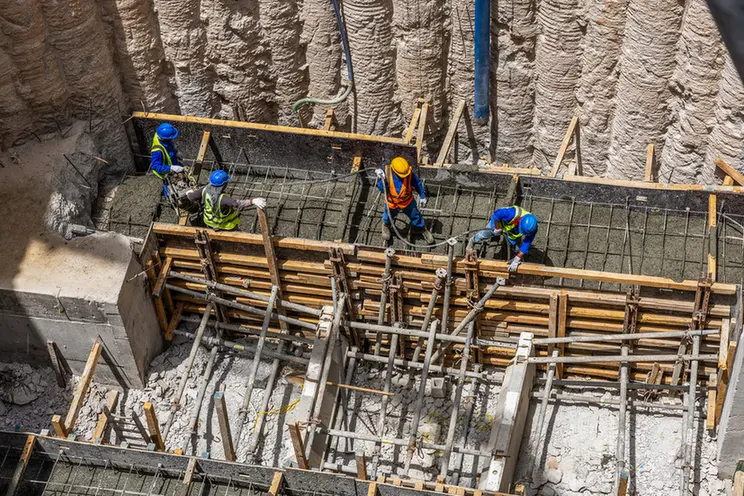 Construction crew in hard hats and safety vests work along wooden formwork and steel reinforcement in a deep trench for concrete.