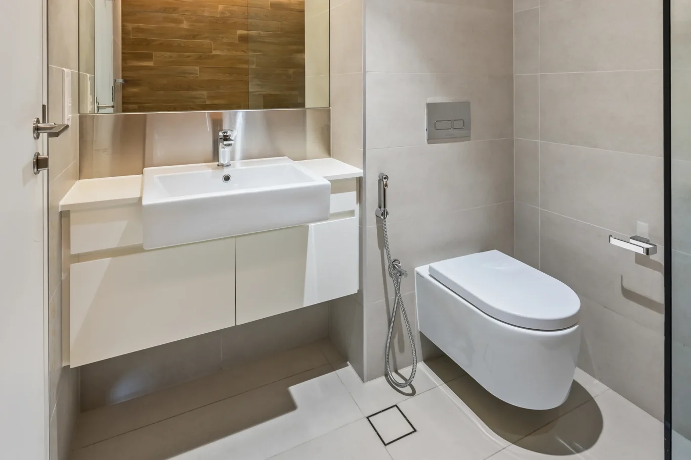 Modern bathroom with a white wall-mounted sink and storage cabinet, large mirror, and beige tiled walls; a wall-mounted toilet with a dual-flush plate and handheld bidet near the toilet visible to the right.