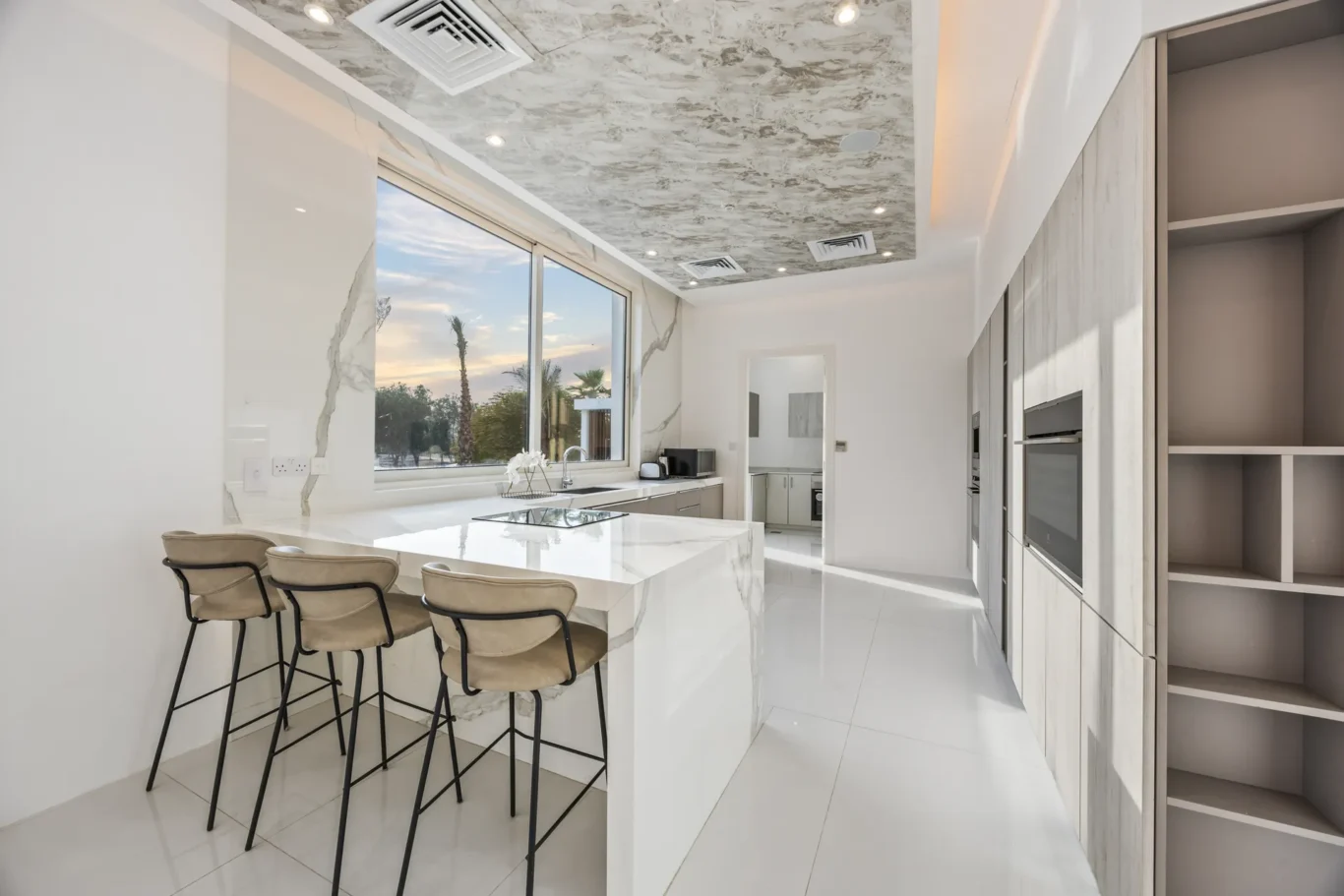 Bright modern kitchen with a white marble island, four beige bar stools, and large windows bringing in natural light. A row of grey wood cabinets lines the right wall with built-in appliances nearby.