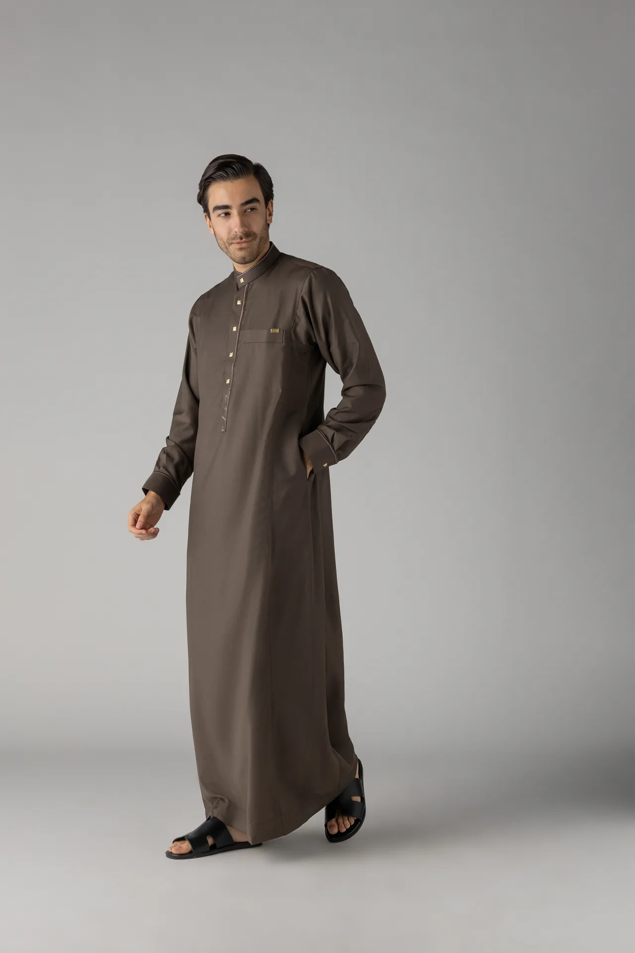 Man wearing a brown traditional kurta with gold buttons, standing with one hand in pocket against a plain gray backdrop.