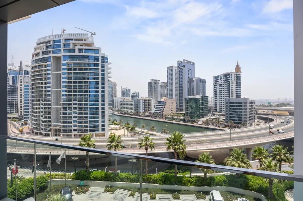 Balcony view of a modern city skyline with blue glass skyscrapers, a waterfront canal, and a looping elevated highway, palm trees in the foreground.