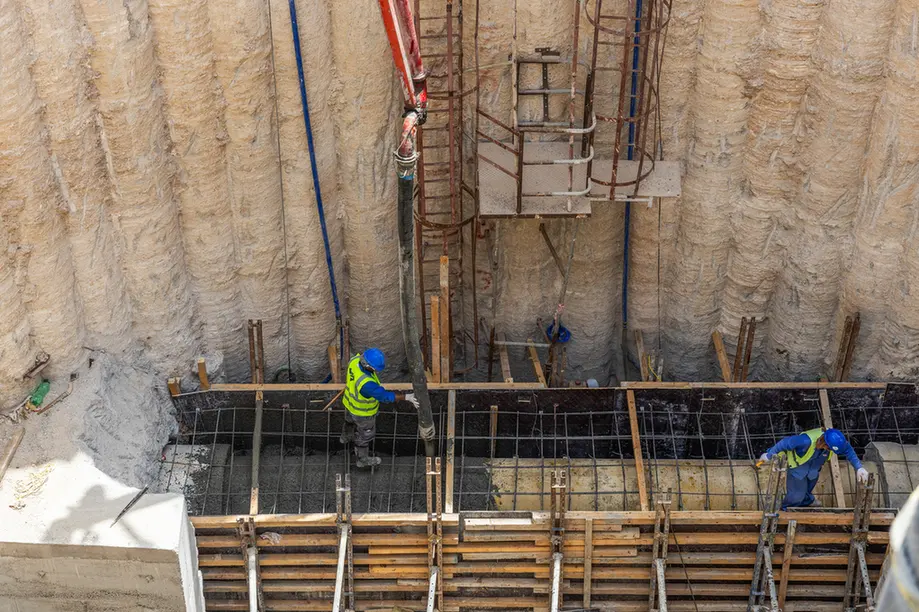 Two construction workers in hard hats and vests pour concrete into a formwork with reinforcing bars in a deep excavation pit.