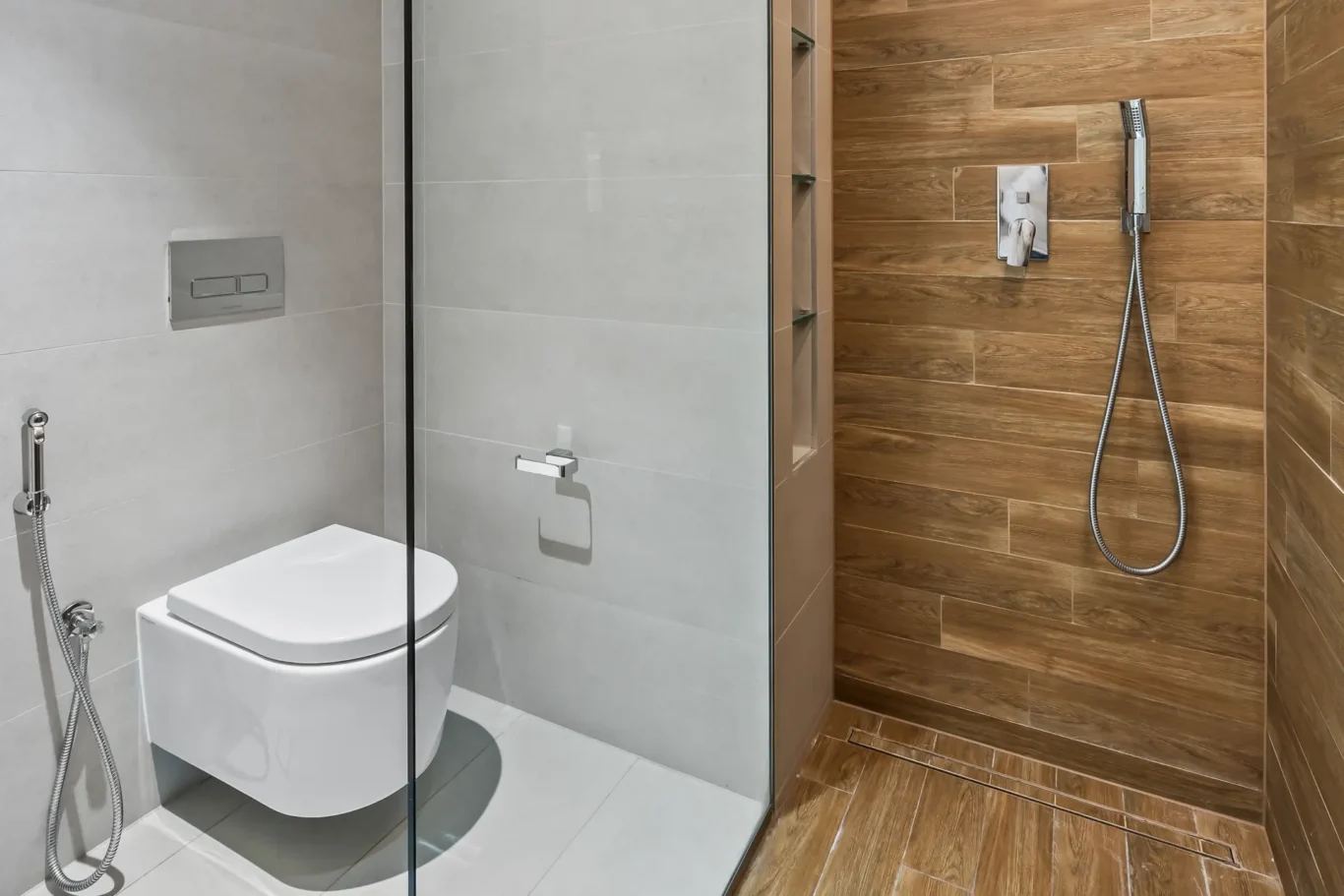Modern bathroom with a wall-mounted toilet, glass partition, and a wood-look tiled shower area with a handheld showerhead on the right.