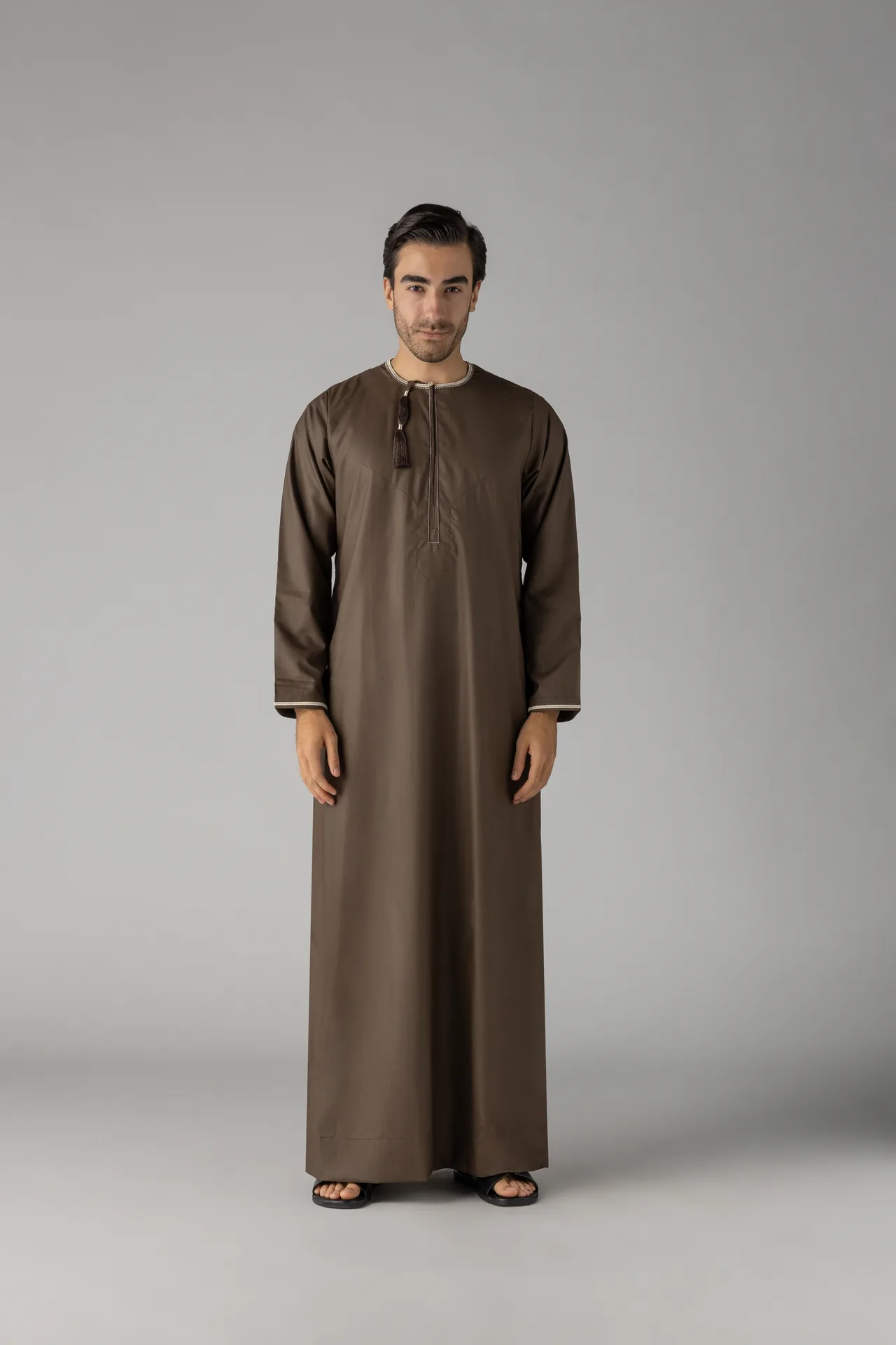Full-length portrait of a man in a brown traditional thobe with a tassel at the neck, standing against a gray studio backdrop, wearing sandals.