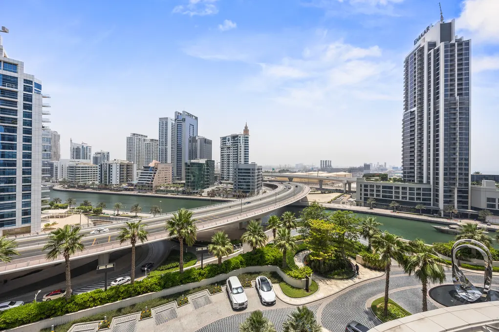 City skyline along a canal with an elevated freeway, palm trees, and modern glass buildings in sunlight.