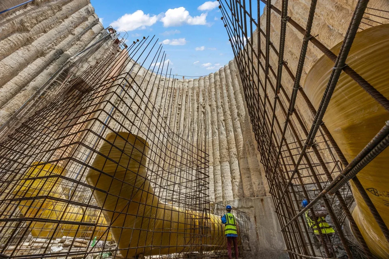 Construction site showing curved concrete wall with vertical steel rebar cages; workers in safety vests and helmets inspect the structure.