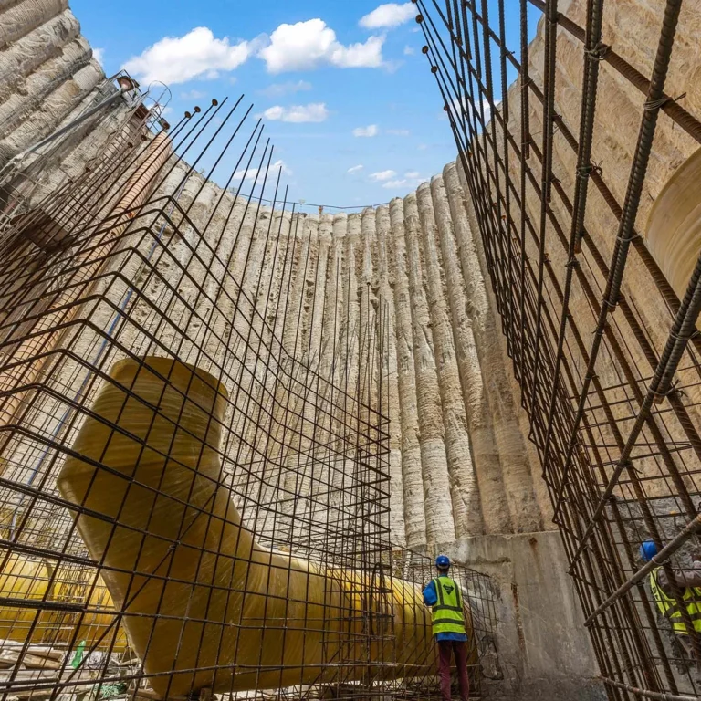 Construction site showing curved concrete wall with vertical steel rebar cages; workers in safety vests and helmets inspect the structure.
