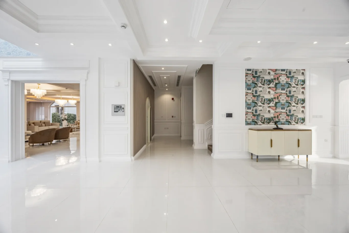 Bright white hotel lobby with high ceilings, glossy floor, and a cream cabinet under a colorful patterned wall panel on the right.