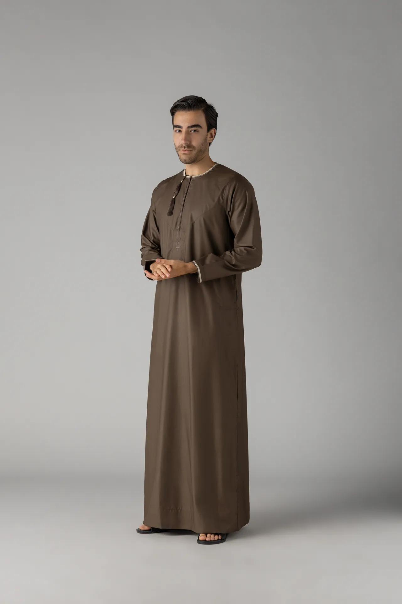 Man wearing a brown thobe stands against a gray backdrop, hands gently clasped in front.