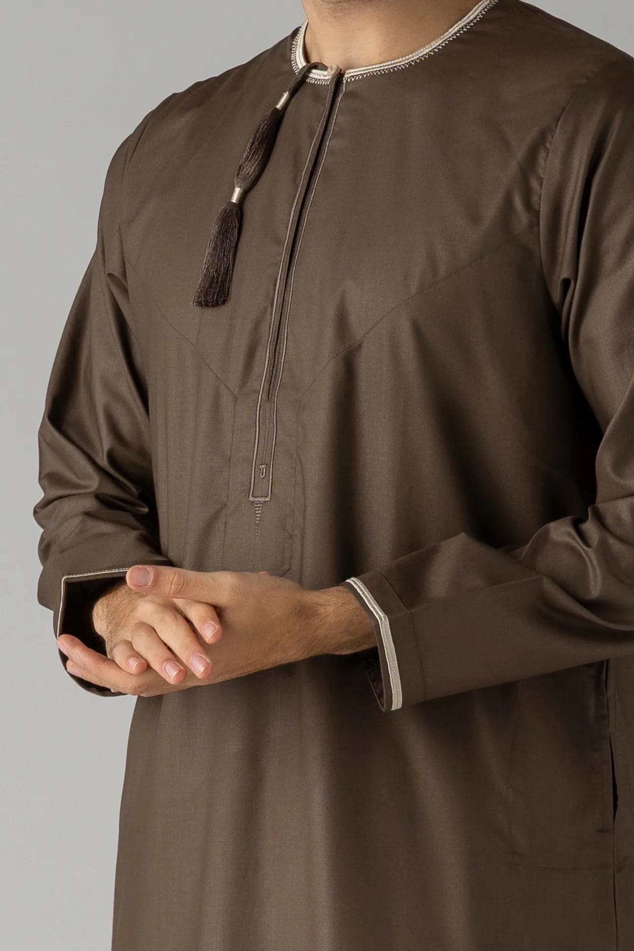 Man wearing a brown thobe with white stitched collar and tassel, holding a smartphone against a gray background.