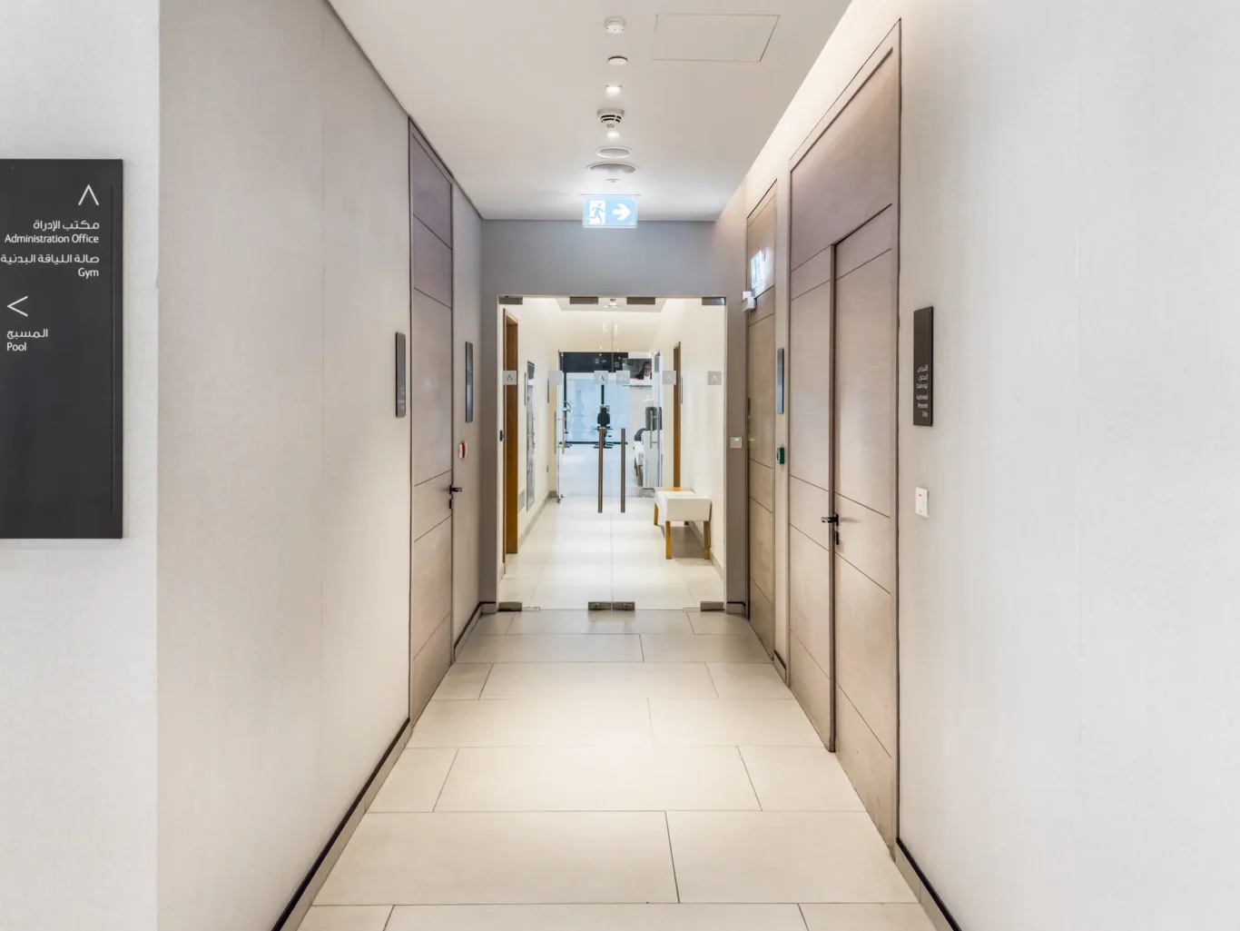 Long hallway in a modern building with closed wooden doors along both walls and an exit sign visible at the far end outside glass doors.