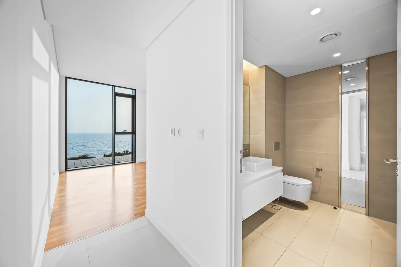 Modern bathroom with beige tile walls, glass shower, and white vanity; hallway opens to a bright living area with ocean view through large windows.