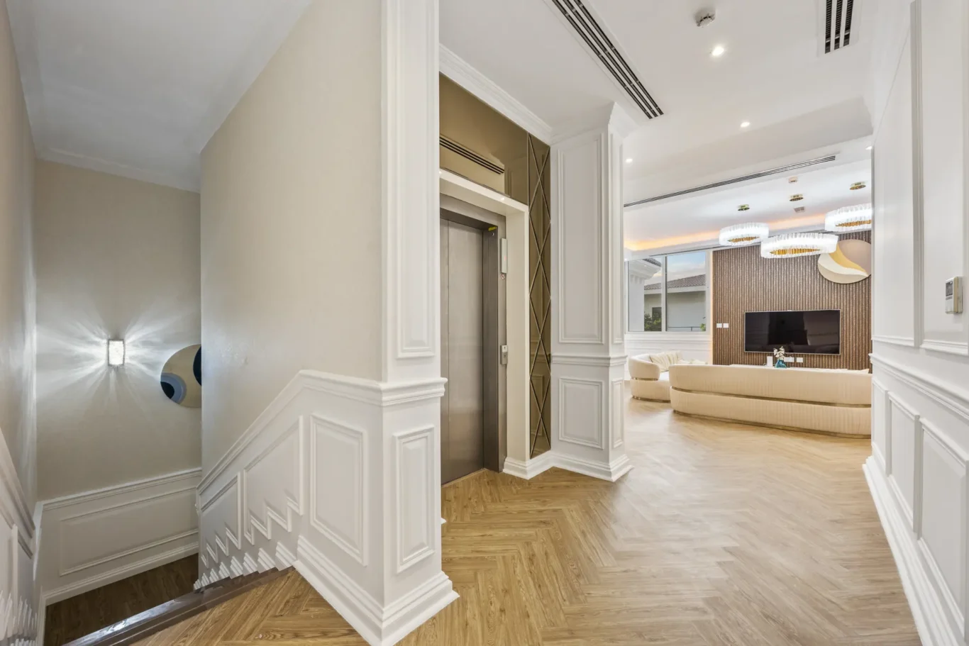 Elegant lobby with an elevator set in white paneled walls, decorative molding, and glass mirrored panels leading to a bright living area with a TV.