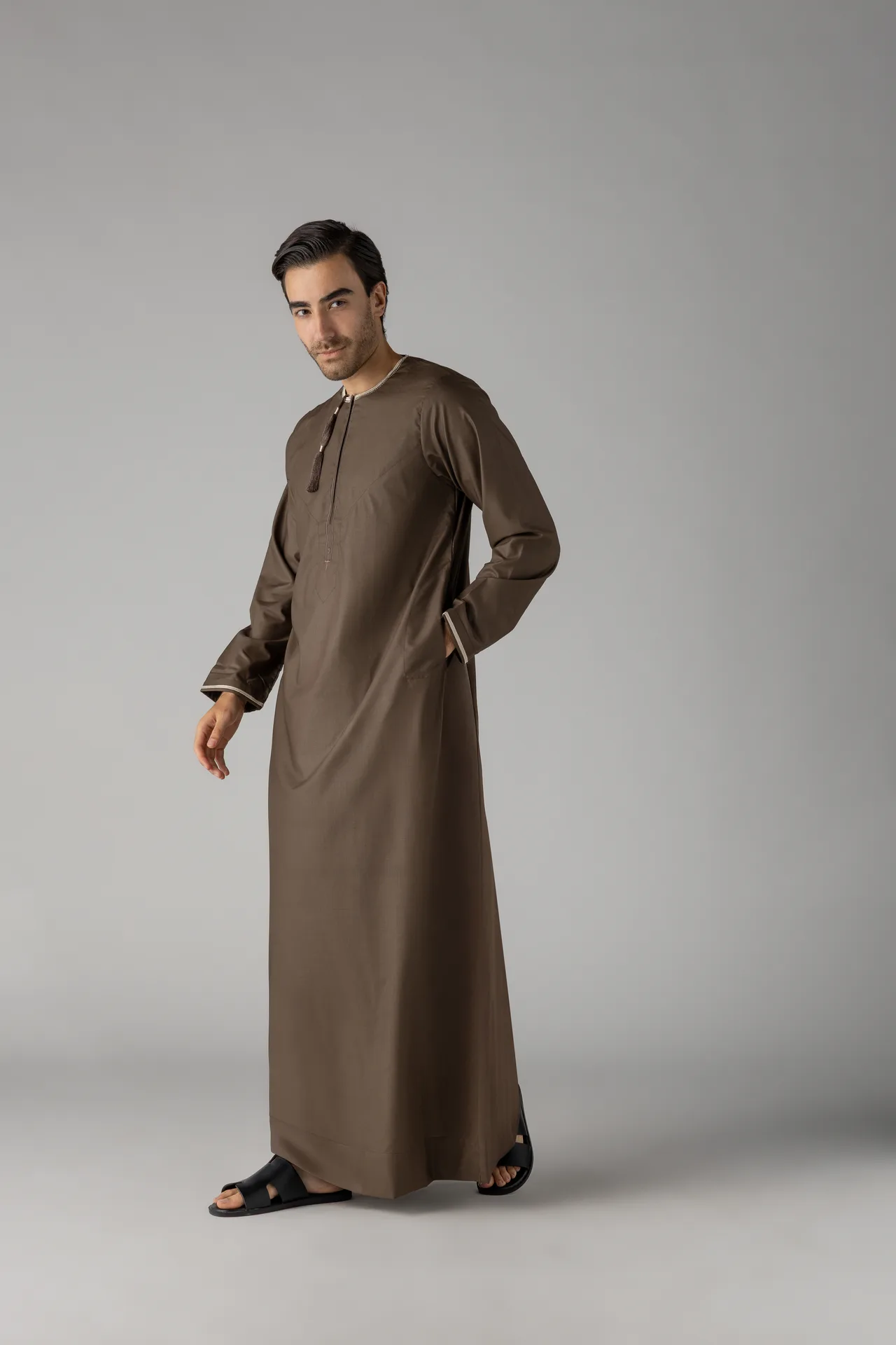 Man in a brown traditional thobe standing against a gray studio backdrop, hands relaxed at his sides, wearing black sandals.