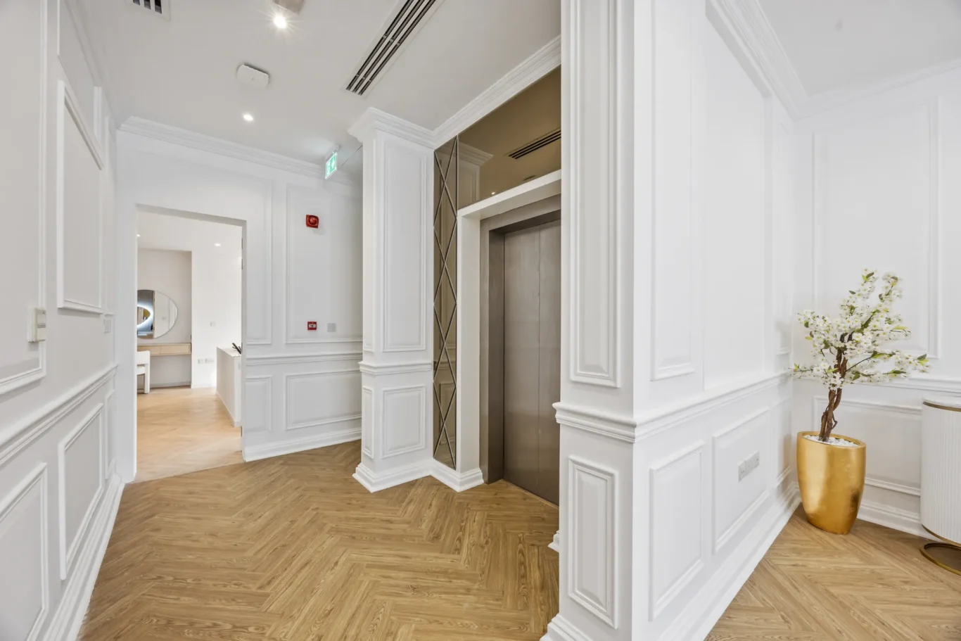 Bright white hallway with paneled walls, an elevator on the right, and a gold vase with white blossoms in the foreground.