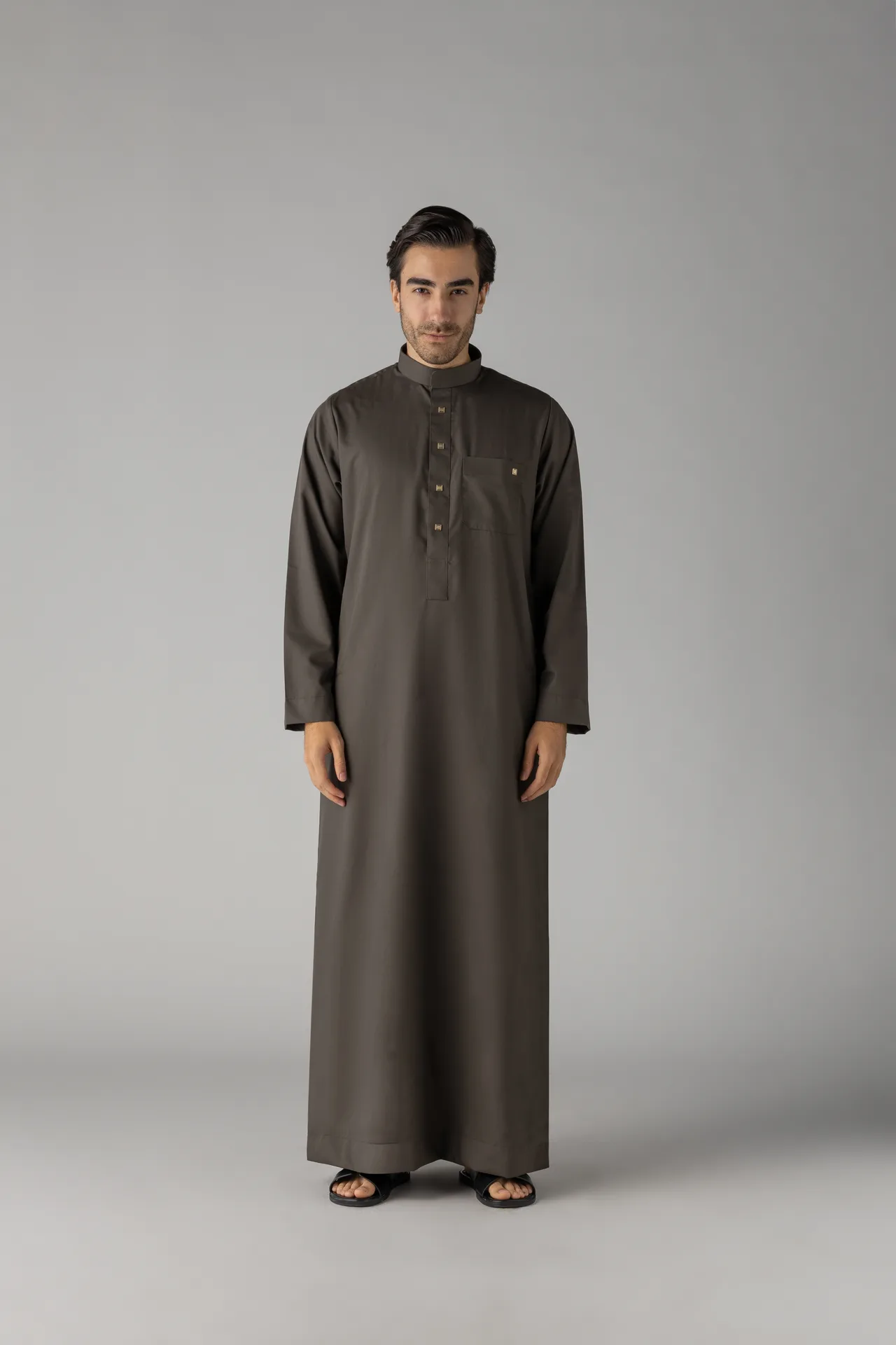 Man wearing a dark gray thobe, standing against a neutral gray background, facing the camera.