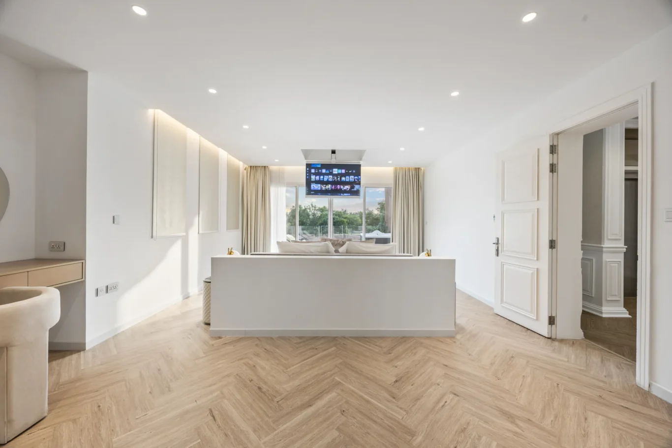 Bright minimalist living room with white walls, pale wood herringbone floor, ceiling spotlights, and a ceiling-mounted TV facing a sofa by large windows.