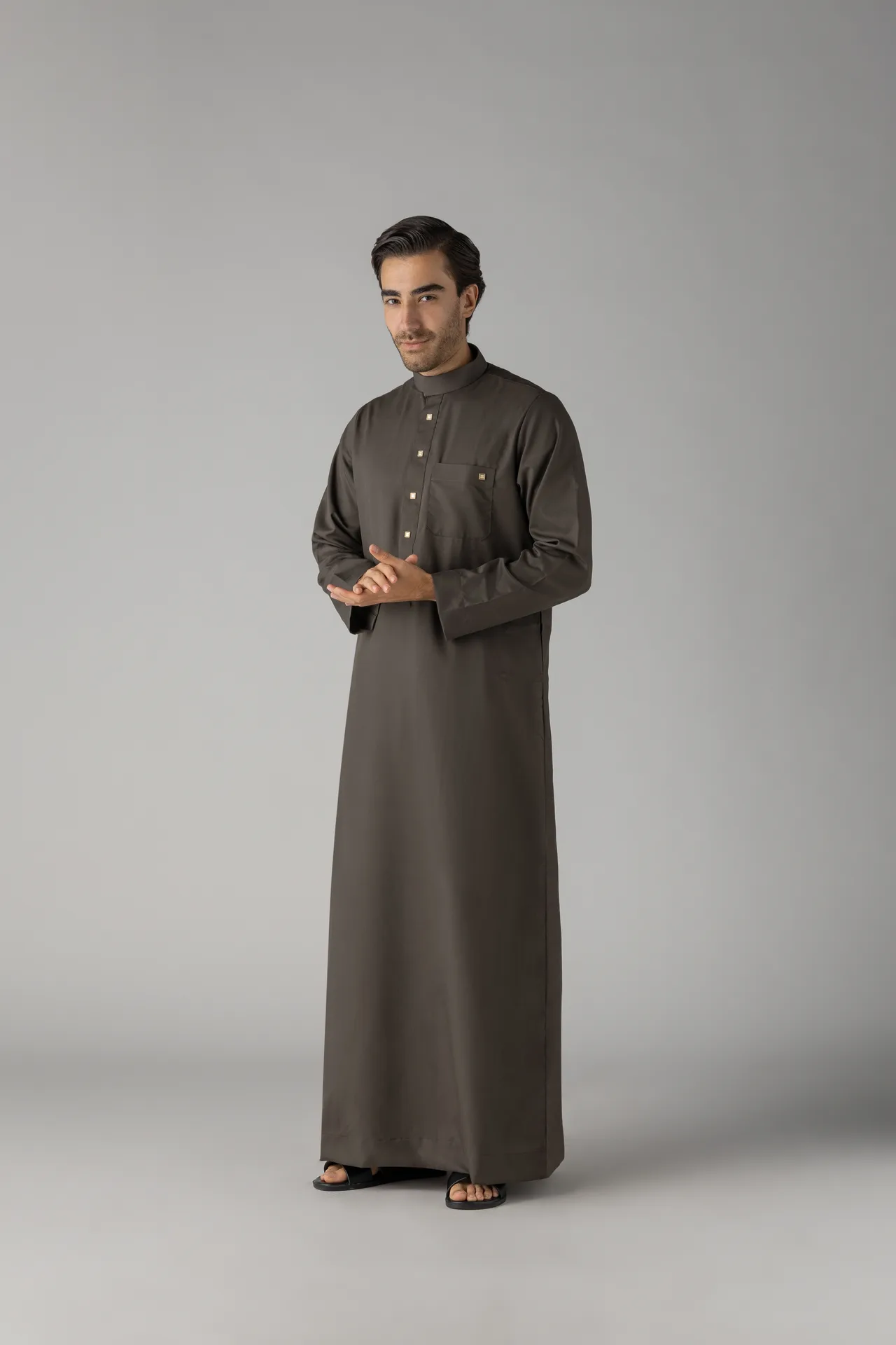 Man wearing a long brown thobe, standing with hands clasped in front, against a gray studio backdrop.