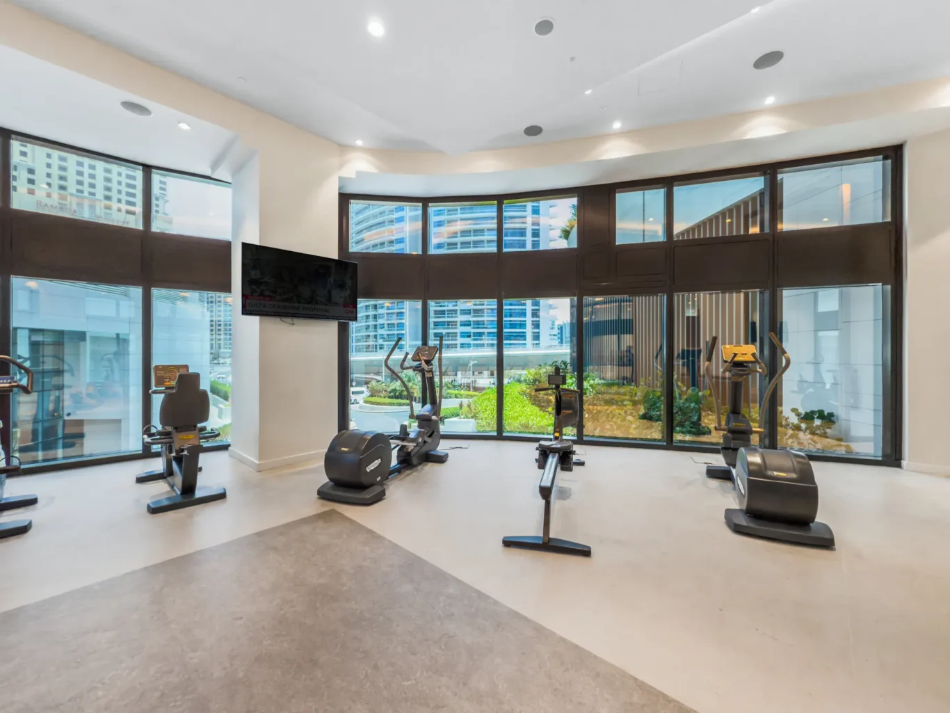 Bright, modern gym with cardio machines (treadmills and bikes) facing curved floor-to-ceiling windows with city views outside.