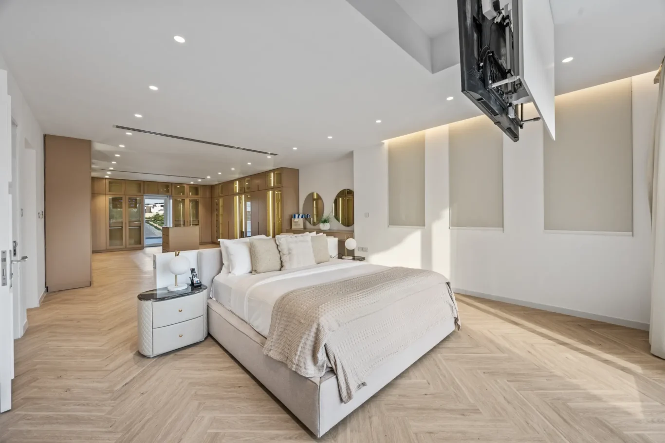 Spacious modern master bedroom with a king bed, beige bedding, round mirrors, and built-in wardrobes along the back wall.