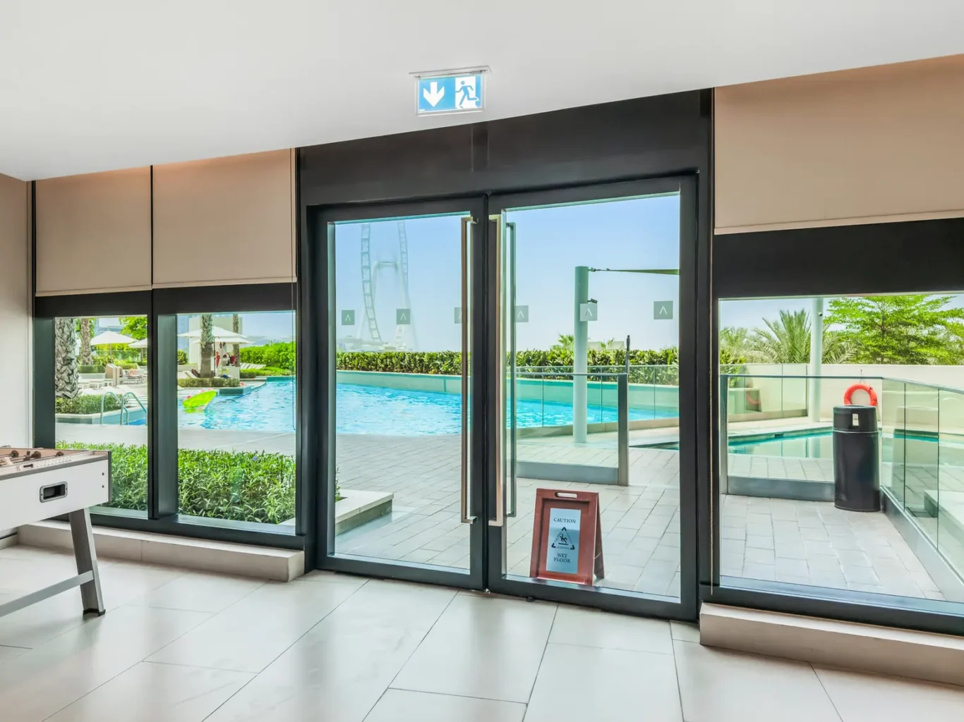 Indoor lobby with large glass doors opening to a sunny pool patio; a foosball table is visible on the left and a wet-floor caution sign near the doorway.