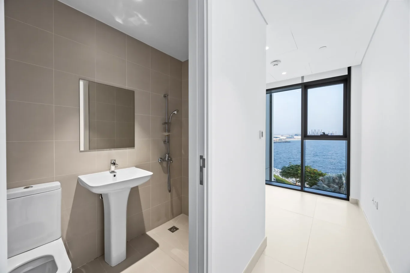 Modern bathroom with beige tiled walls, pedestal sink, toilet, and shower; open doorway to a bright room with a large window overlooking the sea.