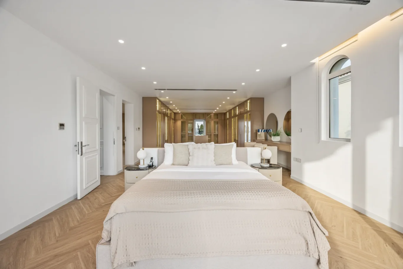 Bright, airy bedroom with a large bed in the foreground, beige bedding, and a mirrored cabinetry wall at the far end.