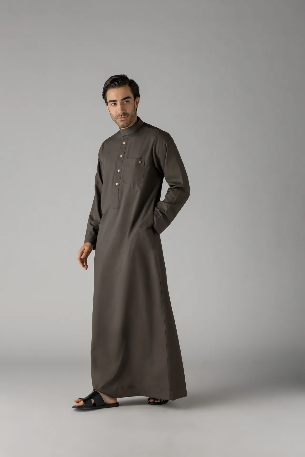 Man wearing a long brown thobe stands in a neutral studio background, one hand in his pocket, wearing black sandals.